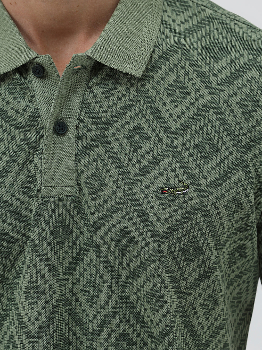 A man wearing a short-sleeve polo shirt with a green and dark gray geometric diamond pattern, paired with dark pants, standing against a light gray studio background.