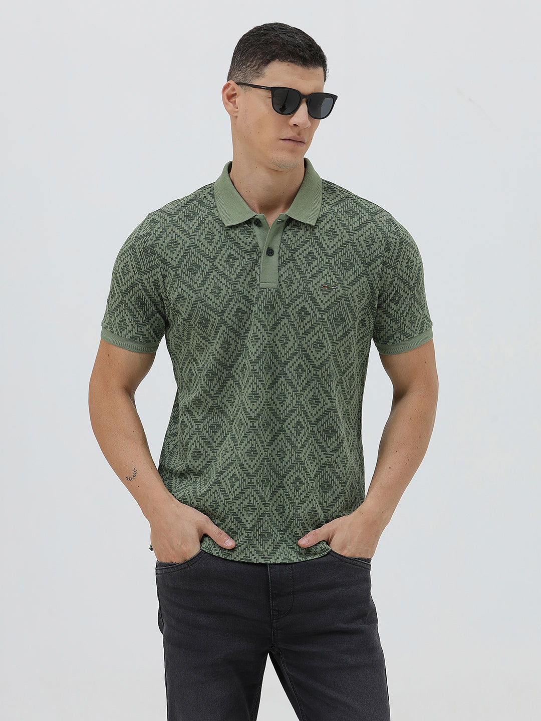 A man wearing a short-sleeve polo shirt with a green and dark gray geometric diamond pattern, paired with dark pants, standing against a light gray studio background.
