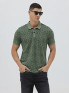 A man wearing a short-sleeve polo shirt with a green and dark gray geometric diamond pattern, paired with dark pants, standing against a light gray studio background.