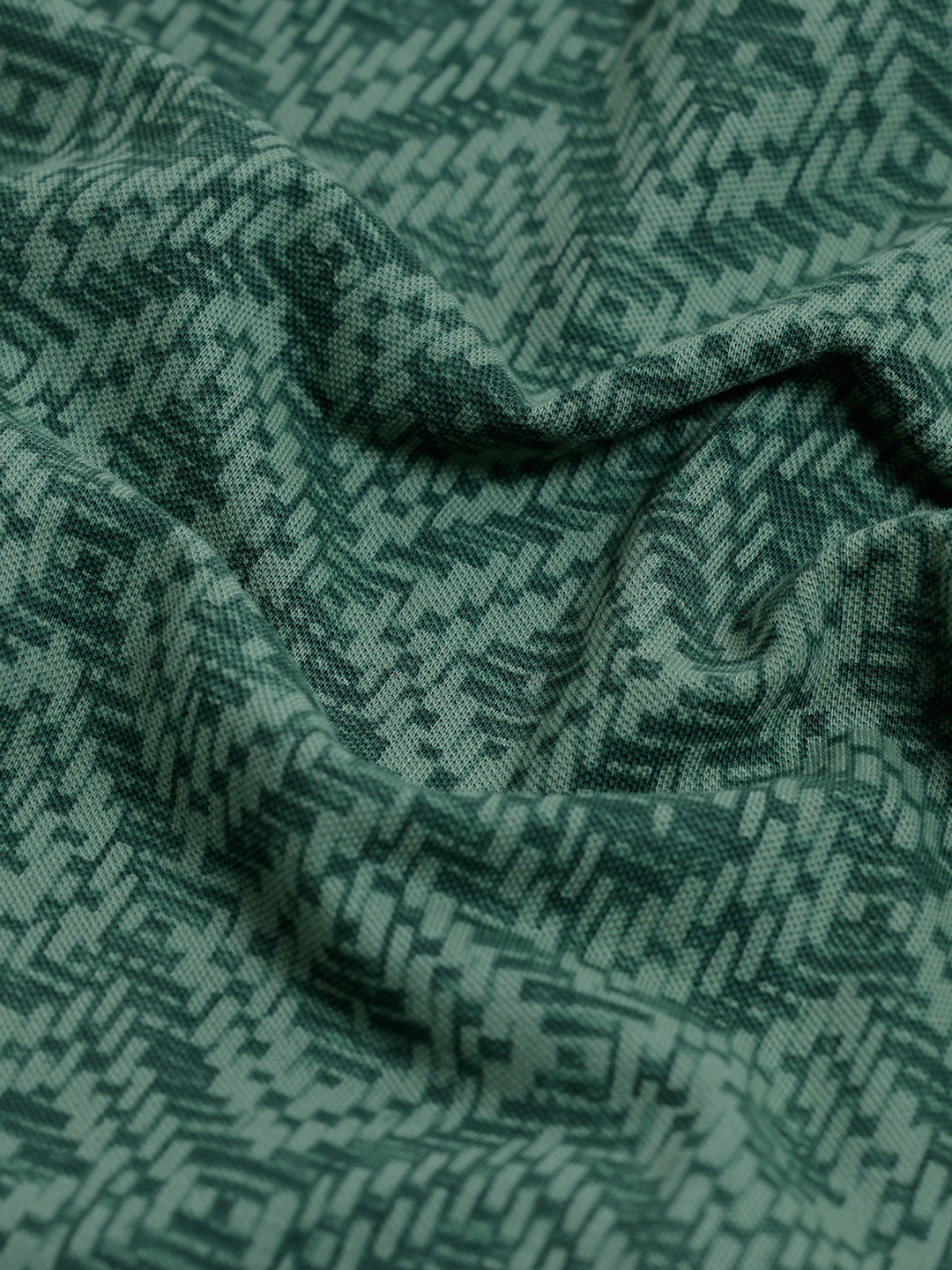A man wearing a short-sleeve polo shirt with a green and dark gray geometric diamond pattern, paired with dark pants, standing against a light gray studio background.