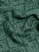 A man wearing a short-sleeve polo shirt with a green and dark gray geometric diamond pattern, paired with dark pants, standing against a light gray studio background.