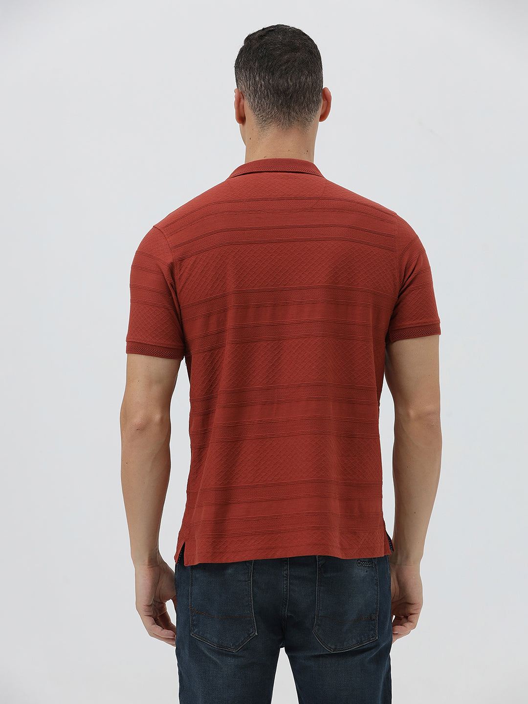 A man wearing a rust-red short-sleeve polo shirt with a subtle textured pattern and dark blue jeans, standing against a light gray studio background.
