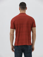 A man wearing a rust-red short-sleeve polo shirt with a subtle textured pattern and dark blue jeans, standing against a light gray studio background.