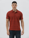 A man wearing a rust-red short-sleeve polo shirt with a subtle textured pattern and dark blue jeans, standing against a light gray studio background.