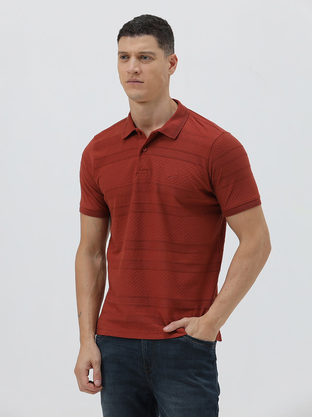 A man wearing a rust-red short-sleeve polo shirt with a subtle textured pattern and dark blue jeans, standing against a light gray studio background.