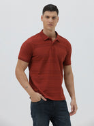 A man wearing a rust-red short-sleeve polo shirt with a subtle textured pattern and dark blue jeans, standing against a light gray studio background.