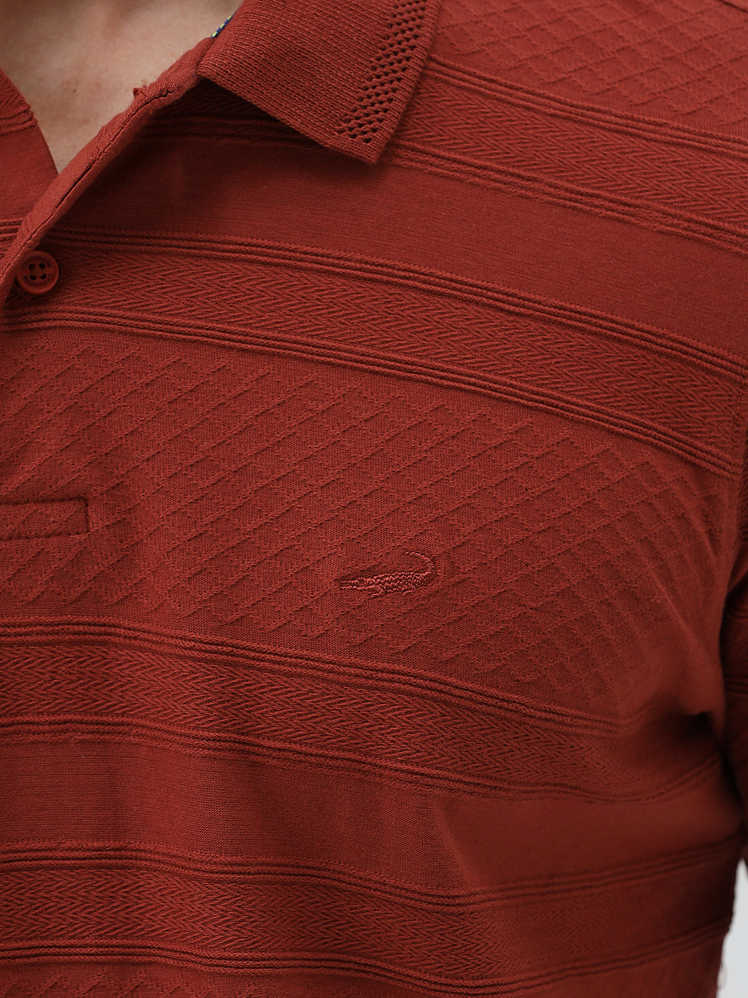 A man wearing a rust-red short-sleeve polo shirt with a subtle textured pattern and dark blue jeans, standing against a light gray studio background.
