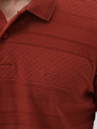 A man wearing a rust-red short-sleeve polo shirt with a subtle textured pattern and dark blue jeans, standing against a light gray studio background.