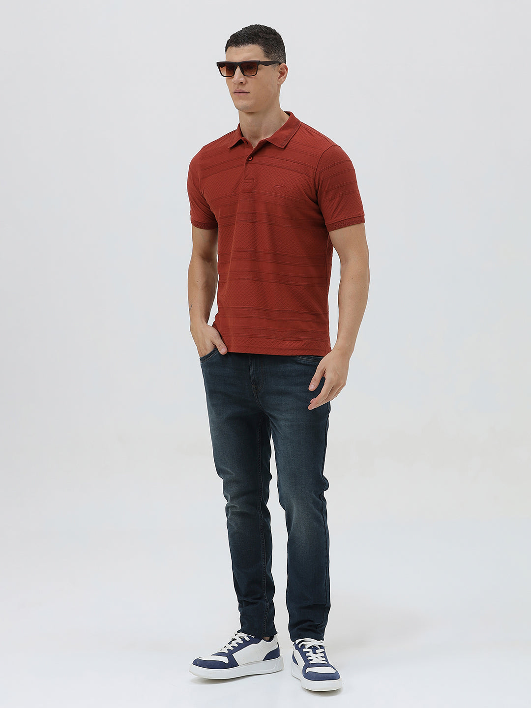 A man wearing a rust-red short-sleeve polo shirt with a subtle textured pattern and dark blue jeans, standing against a light gray studio background.