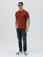 A man wearing a rust-red short-sleeve polo shirt with a subtle textured pattern and dark blue jeans, standing against a light gray studio background.