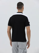 A man modeling a black short-sleeve polo shirt with white shoulder panels and white striped trim on the collar and sleeve cuffs, paired with light gray cargo pants against a plain light gray background.