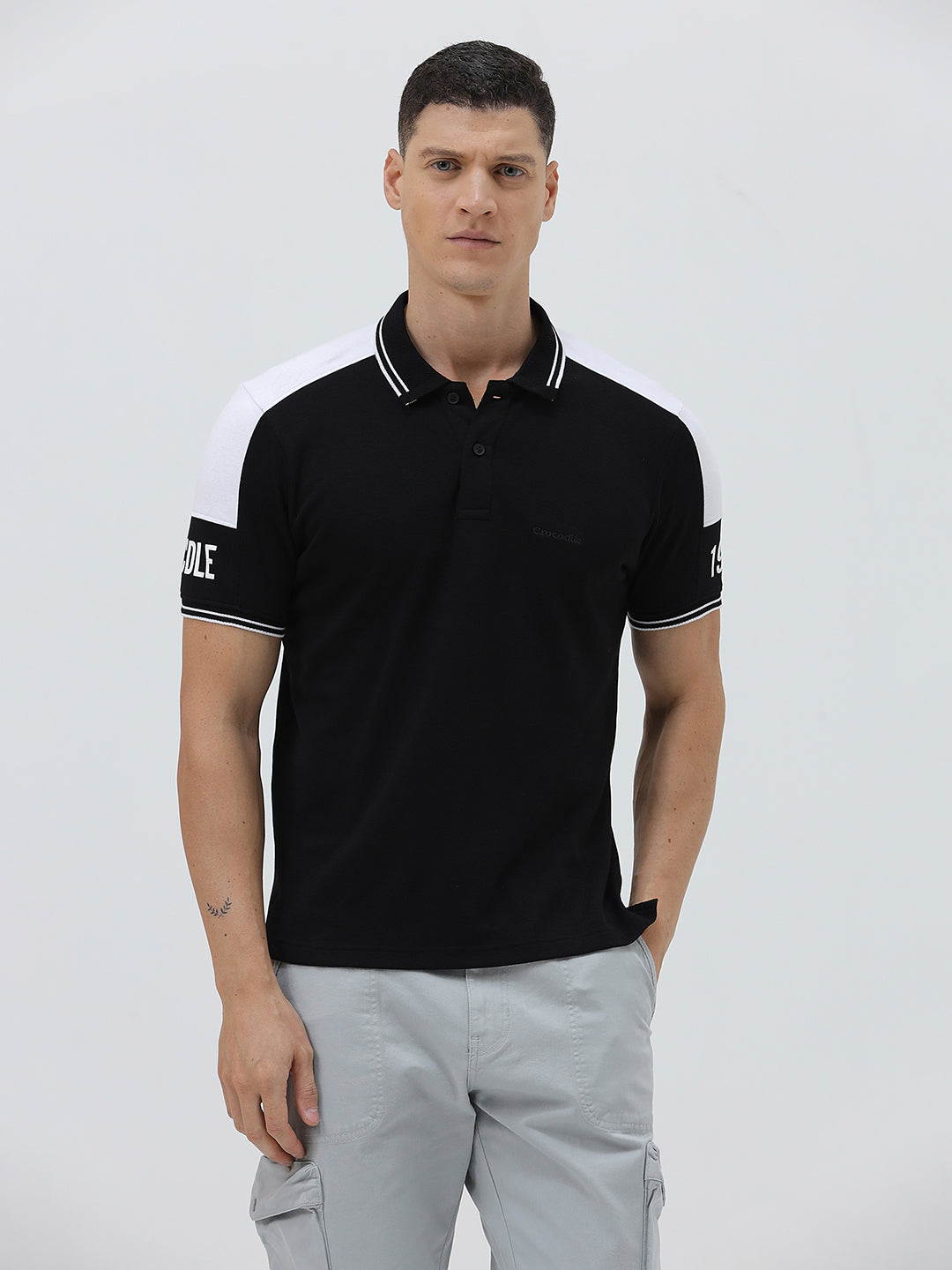 A man modeling a black short-sleeve polo shirt with white shoulder panels and white striped trim on the collar and sleeve cuffs, paired with light gray cargo pants against a plain light gray background.