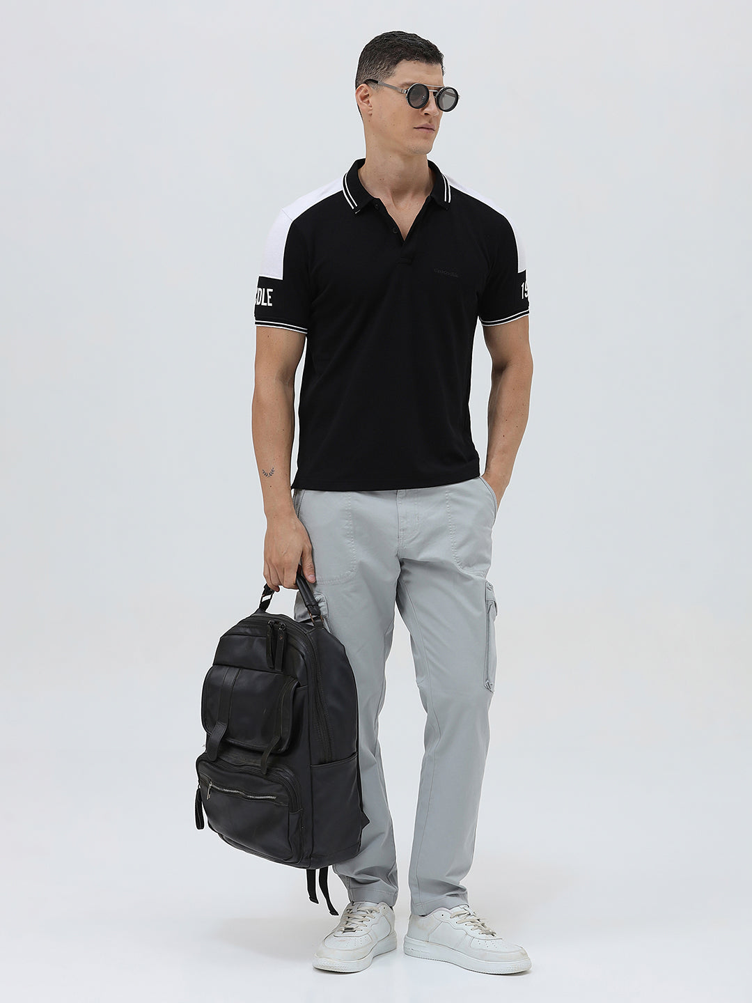A man modeling a black short-sleeve polo shirt with white shoulder panels and white striped trim on the collar and sleeve cuffs, paired with light gray cargo pants against a plain light gray background.