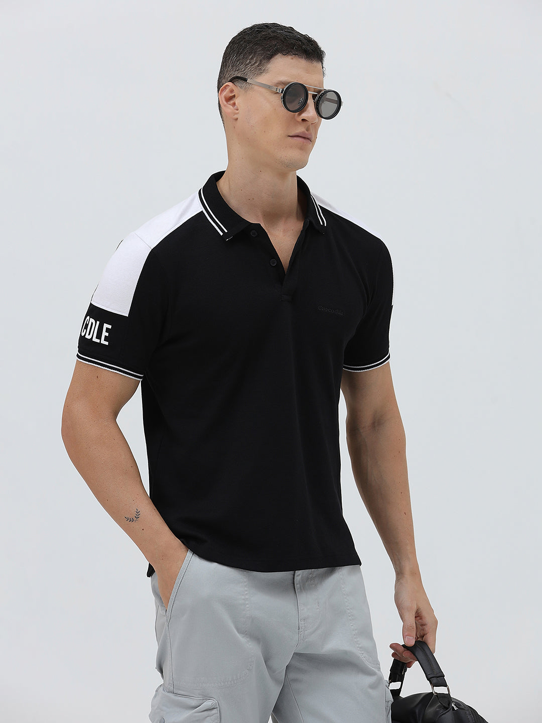 A man modeling a black short-sleeve polo shirt with white shoulder panels and white striped trim on the collar and sleeve cuffs, paired with light gray cargo pants against a plain light gray background.