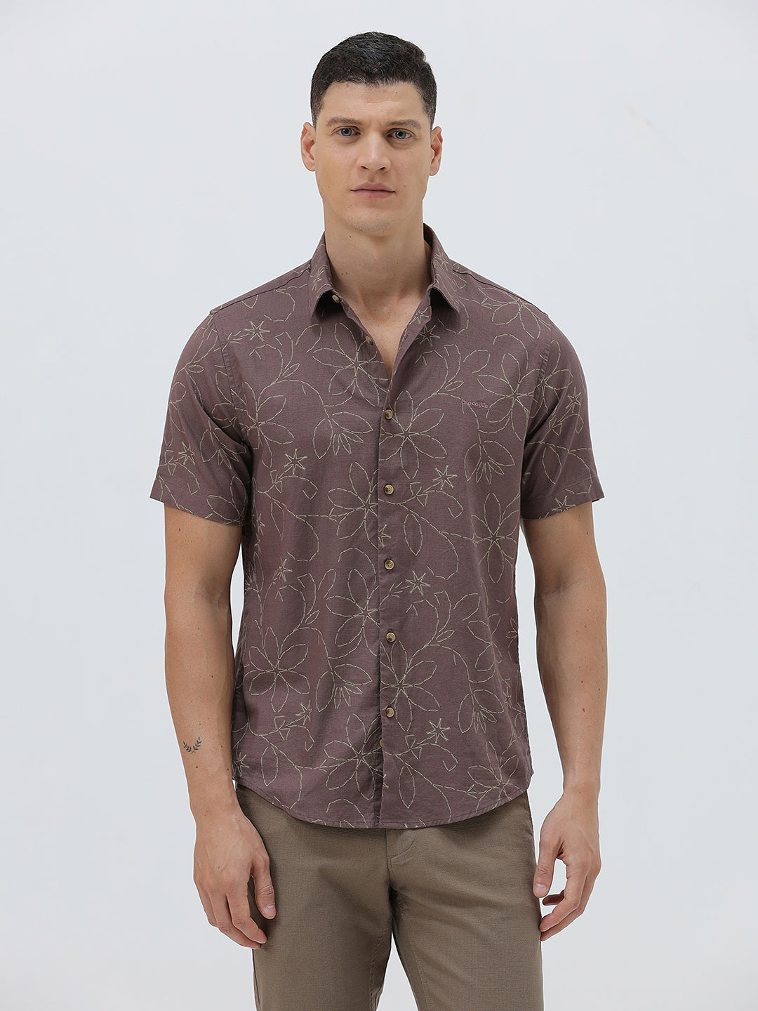 A man wearing a short-sleeve button-up shirt in a muted brownish-purple color with a light beige floral pattern, paired with khaki pants, against a plain light gray background.