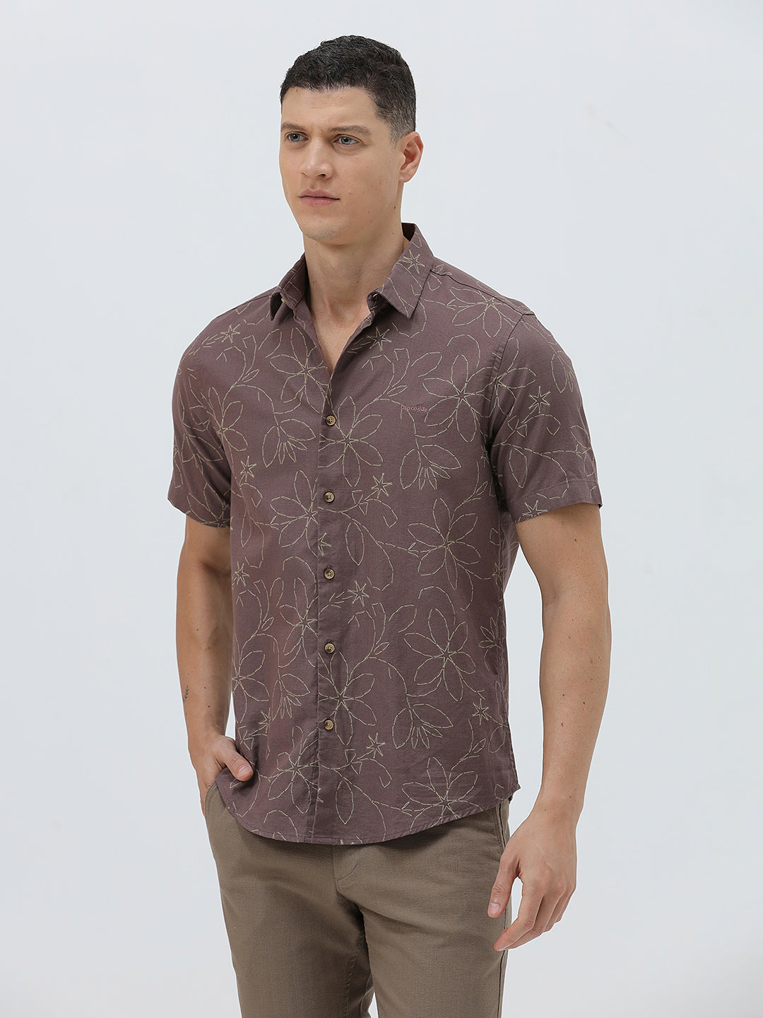 A man wearing a short-sleeve button-up shirt in a muted brownish-purple color with a light beige floral pattern, paired with khaki pants, against a plain light gray background.