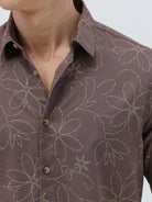 A man wearing a short-sleeve button-up shirt in a muted brownish-purple color with a light beige floral pattern, paired with khaki pants, against a plain light gray background.
