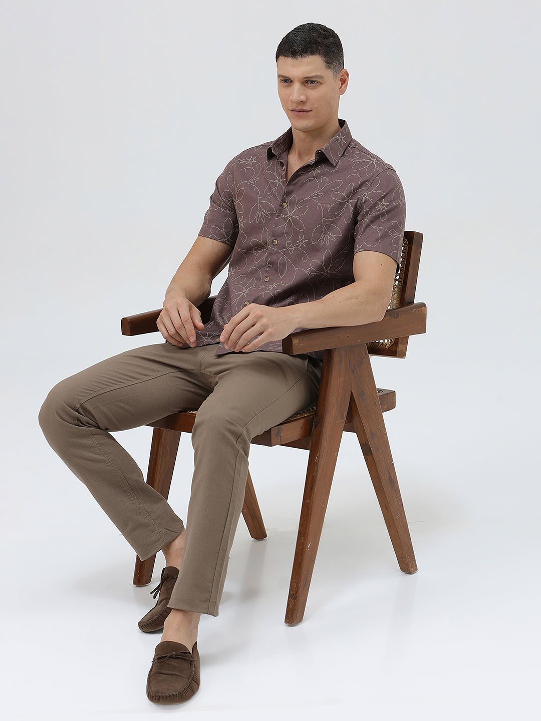 A man wearing a short-sleeve button-up shirt in a muted brownish-purple color with a light beige floral pattern, paired with khaki pants, against a plain light gray background.