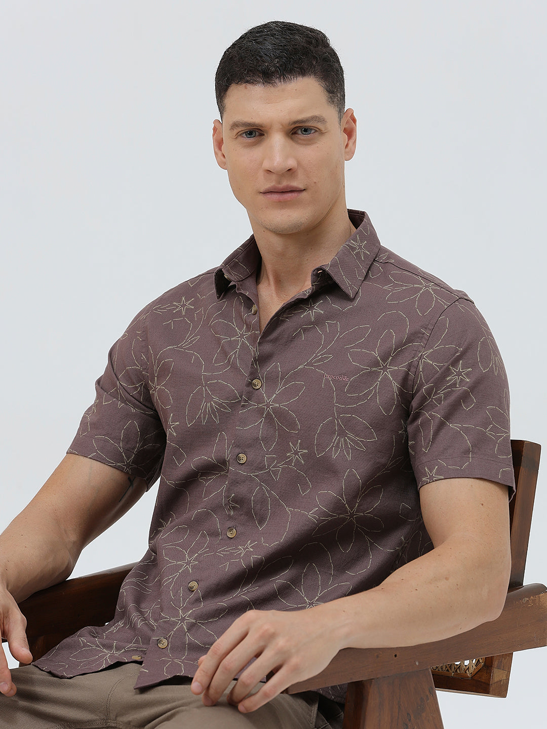 A man wearing a short-sleeve button-up shirt in a muted brownish-purple color with a light beige floral pattern, paired with khaki pants, against a plain light gray background.
