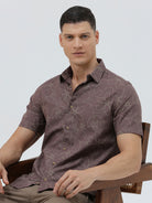 A man wearing a short-sleeve button-up shirt in a muted brownish-purple color with a light beige floral pattern, paired with khaki pants, against a plain light gray background.