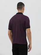 A man wearing a short-sleeve polo shirt in dark plum purple with a black collar and black sleeve trim, paired with black pants, against a plain light gray background.