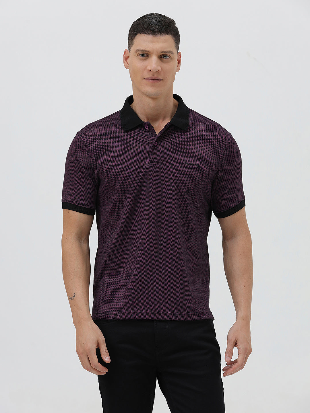 A man wearing a short-sleeve polo shirt in dark plum purple with a black collar and black sleeve trim, paired with black pants, against a plain light gray background.