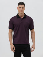 A man wearing a short-sleeve polo shirt in dark plum purple with a black collar and black sleeve trim, paired with black pants, against a plain light gray background.
