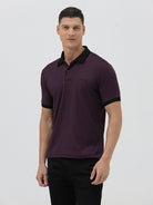 A man wearing a short-sleeve polo shirt in dark plum purple with a black collar and black sleeve trim, paired with black pants, against a plain light gray background.