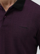 A man wearing a short-sleeve polo shirt in dark plum purple with a black collar and black sleeve trim, paired with black pants, against a plain light gray background.