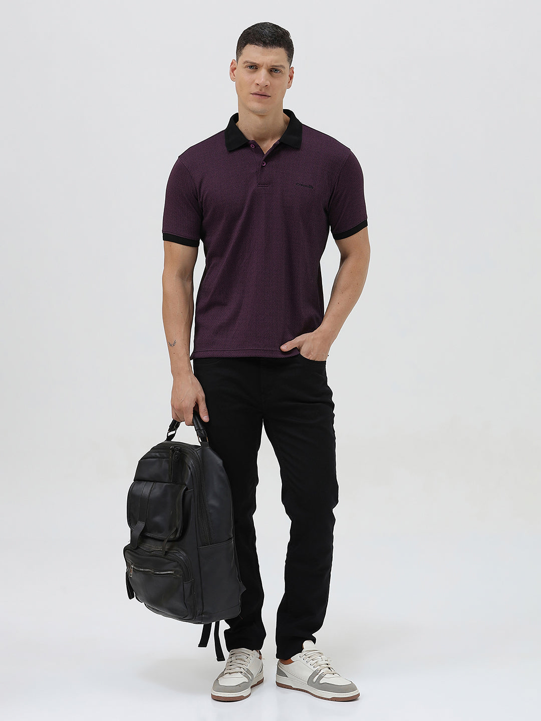 A man wearing a short-sleeve polo shirt in dark plum purple with a black collar and black sleeve trim, paired with black pants, against a plain light gray background.