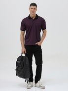 A man wearing a short-sleeve polo shirt in dark plum purple with a black collar and black sleeve trim, paired with black pants, against a plain light gray background.