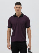 A man wearing a short-sleeve polo shirt in dark plum purple with a black collar and black sleeve trim, paired with black pants, against a plain light gray background.