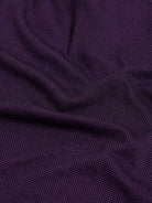 A man wearing a short-sleeve polo shirt in dark plum purple with a black collar and black sleeve trim, paired with black pants, against a plain light gray background.