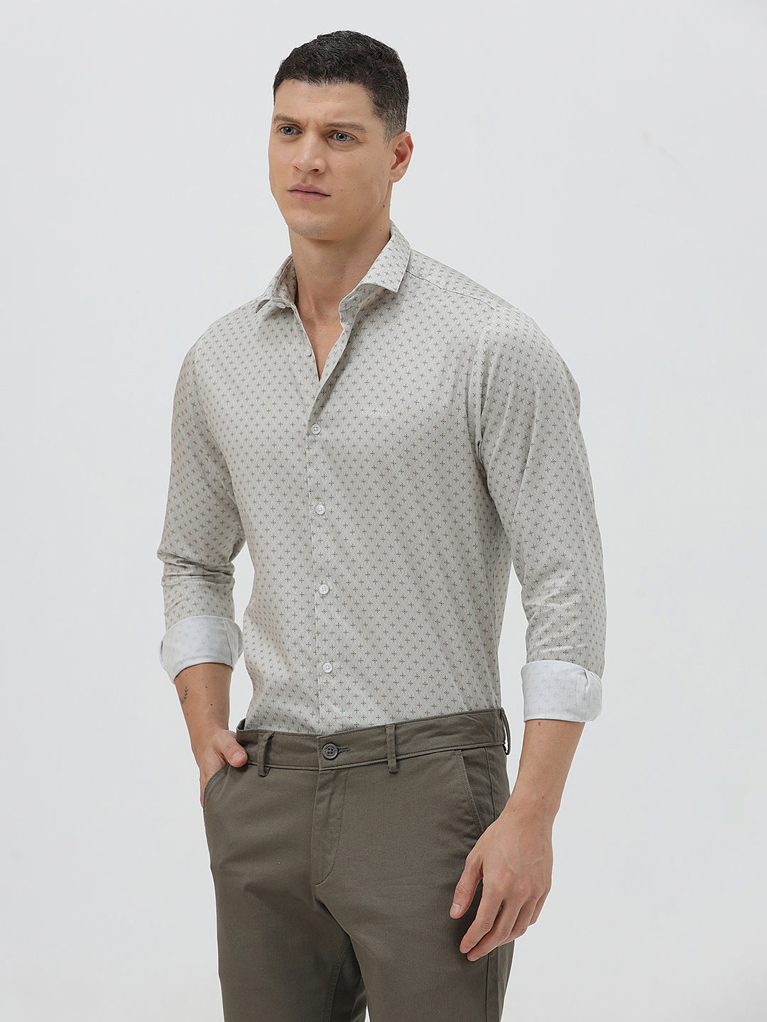 A man wearing a light gray long-sleeve button-down shirt with a small repeating geometric pattern, paired with olive green trousers, against a plain light gray background.