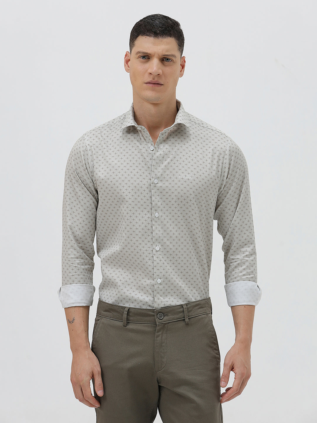 A man wearing a light gray long-sleeve button-down shirt with a small repeating geometric pattern, paired with olive green trousers, against a plain light gray background.