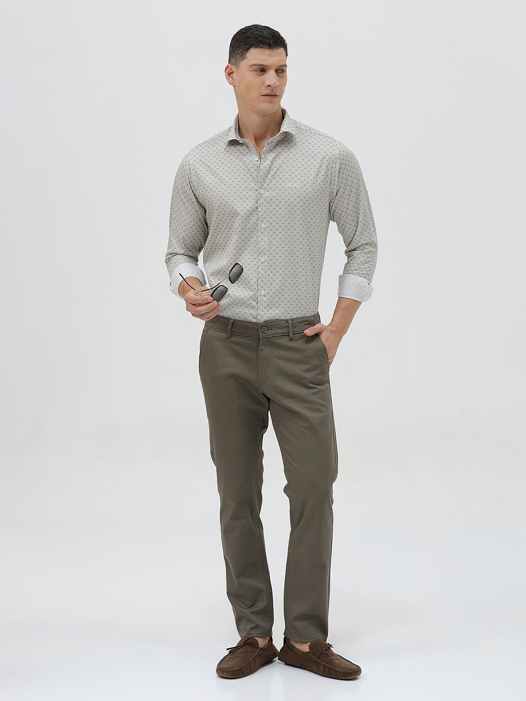 A man wearing a light gray long-sleeve button-down shirt with a small repeating geometric pattern, paired with olive green trousers, against a plain light gray background.