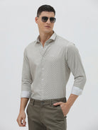 A man wearing a light gray long-sleeve button-down shirt with a small repeating geometric pattern, paired with olive green trousers, against a plain light gray background.