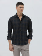 A man wearing a dark charcoal gray long-sleeve button-down shirt with a subtle grid pattern, paired with light gray trousers, against a plain light gray background.