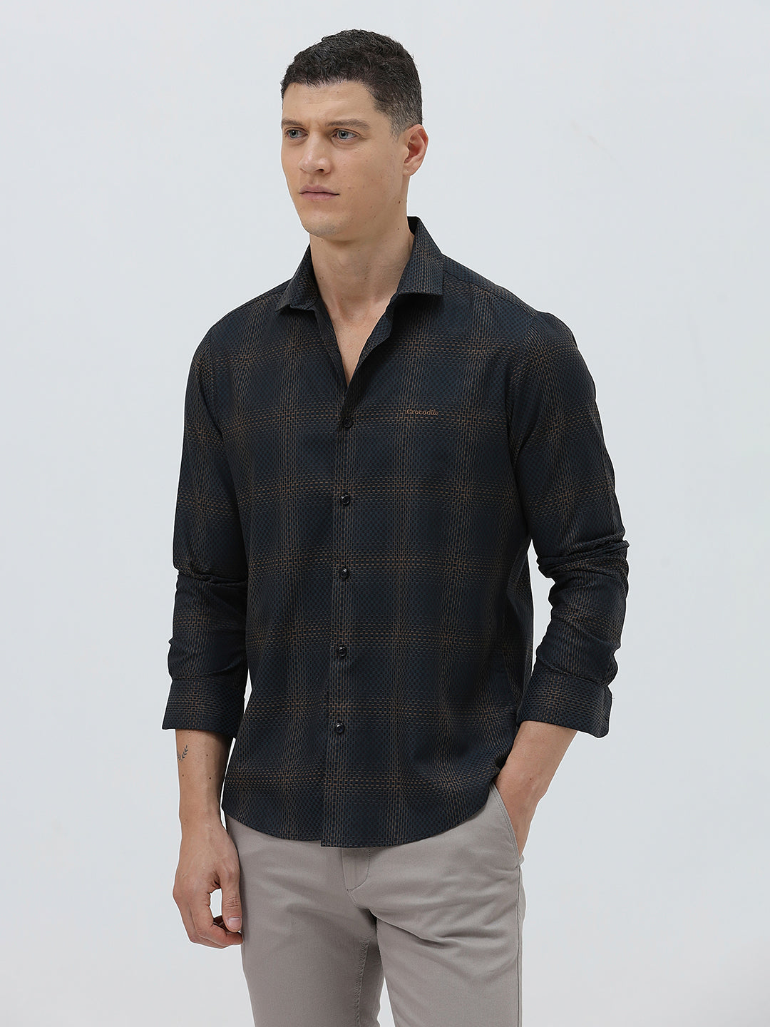 A man wearing a dark charcoal gray long-sleeve button-down shirt with a subtle grid pattern, paired with light gray trousers, against a plain light gray background.
