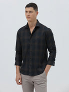 A man wearing a dark charcoal gray long-sleeve button-down shirt with a subtle grid pattern, paired with light gray trousers, against a plain light gray background.