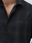 A man wearing a dark charcoal gray long-sleeve button-down shirt with a subtle grid pattern, paired with light gray trousers, against a plain light gray background.