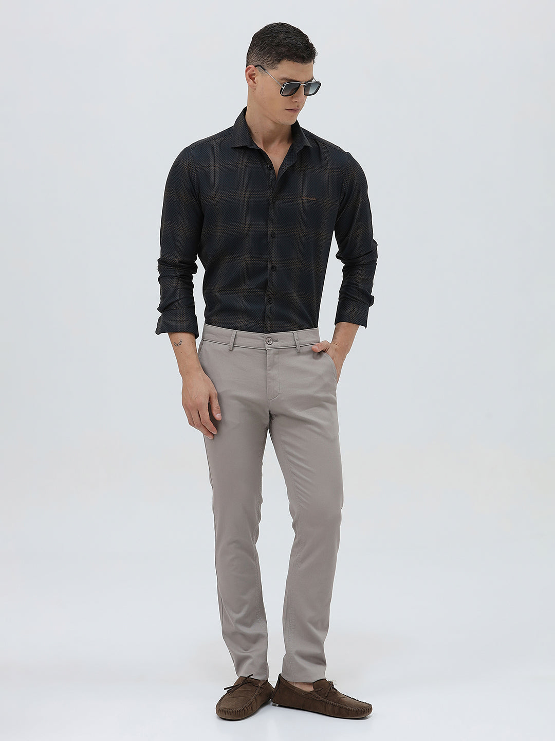 A man wearing a dark charcoal gray long-sleeve button-down shirt with a subtle grid pattern, paired with light gray trousers, against a plain light gray background.