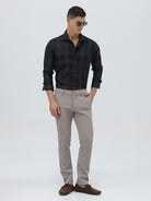 A man wearing a dark charcoal gray long-sleeve button-down shirt with a subtle grid pattern, paired with light gray trousers, against a plain light gray background.