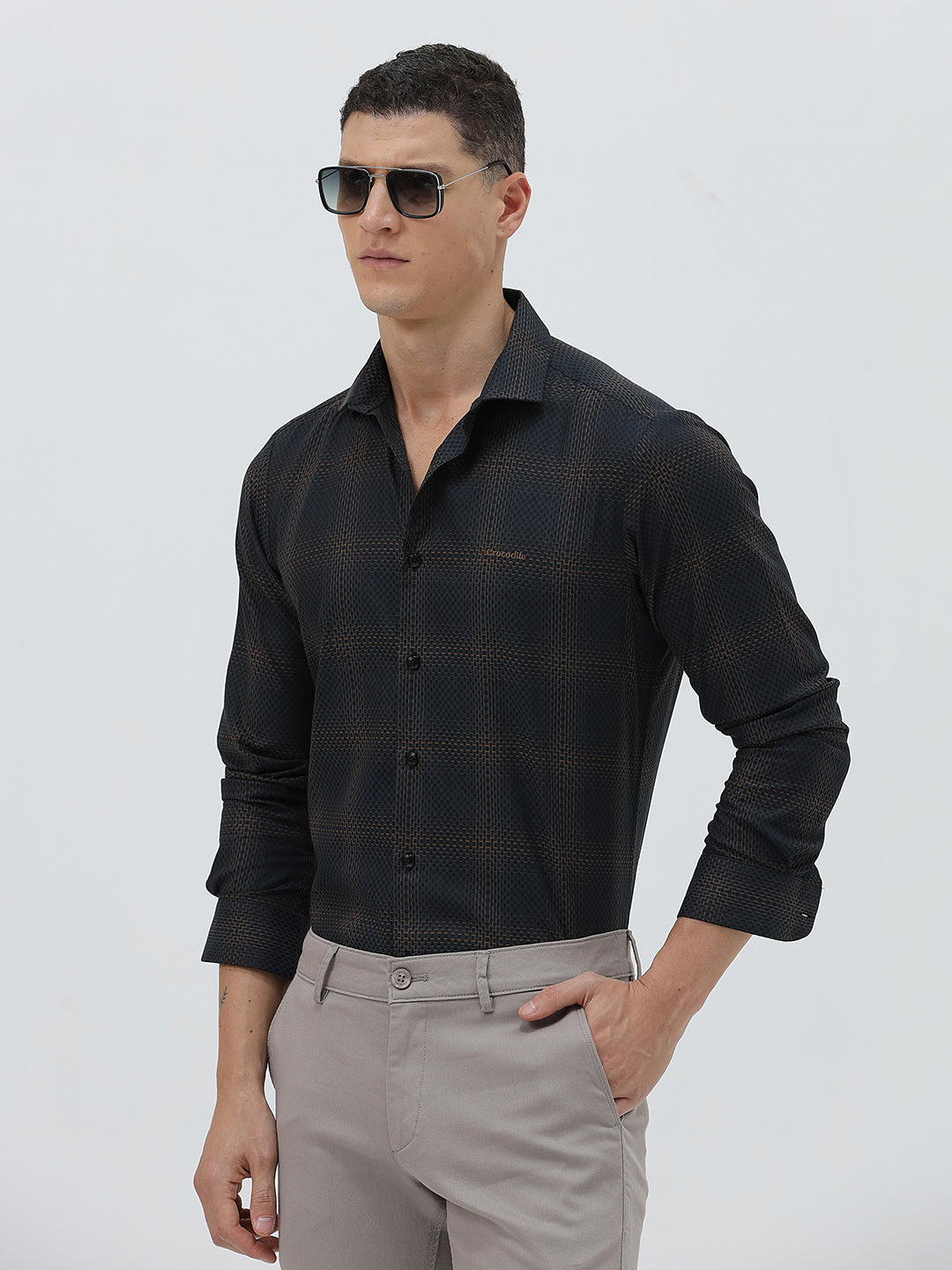 A man wearing a dark charcoal gray long-sleeve button-down shirt with a subtle grid pattern, paired with light gray trousers, against a plain light gray background.
