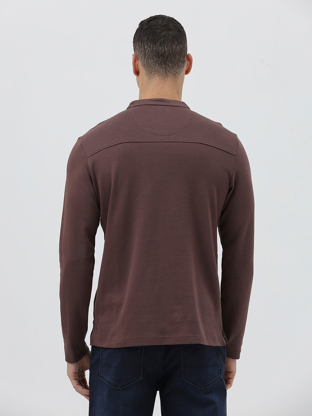 A man wearing a brown long-sleeve henley shirt with a small embroidered logo on the left chest, paired with dark blue jeans, standing against a light gray background.