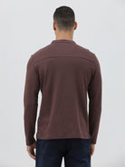 A man wearing a brown long-sleeve henley shirt with a small embroidered logo on the left chest, paired with dark blue jeans, standing against a light gray background.