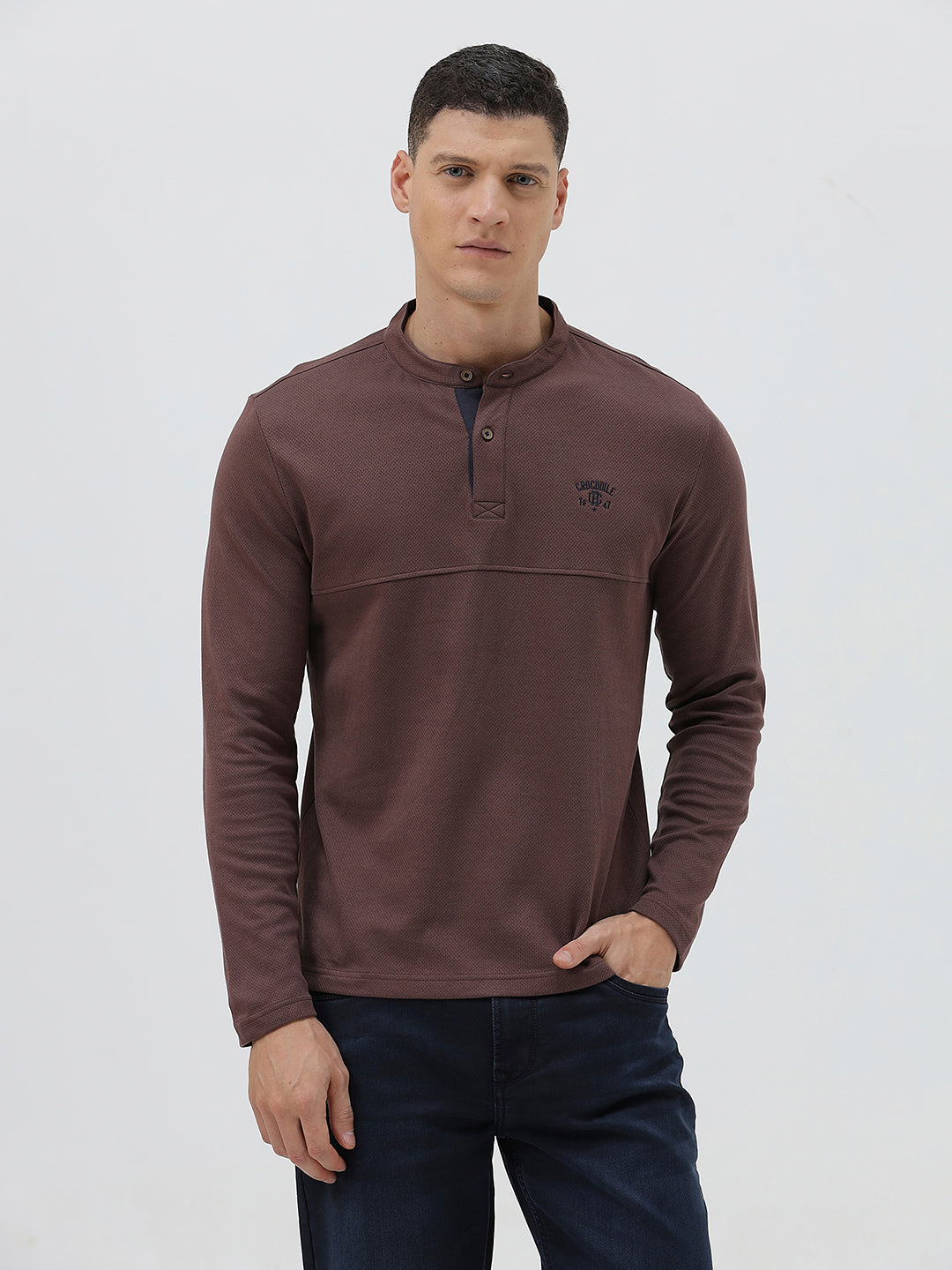 A man wearing a brown long-sleeve henley shirt with a small embroidered logo on the left chest, paired with dark blue jeans, standing against a light gray background.