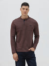 A man wearing a brown long-sleeve henley shirt with a small embroidered logo on the left chest, paired with dark blue jeans, standing against a light gray background.
