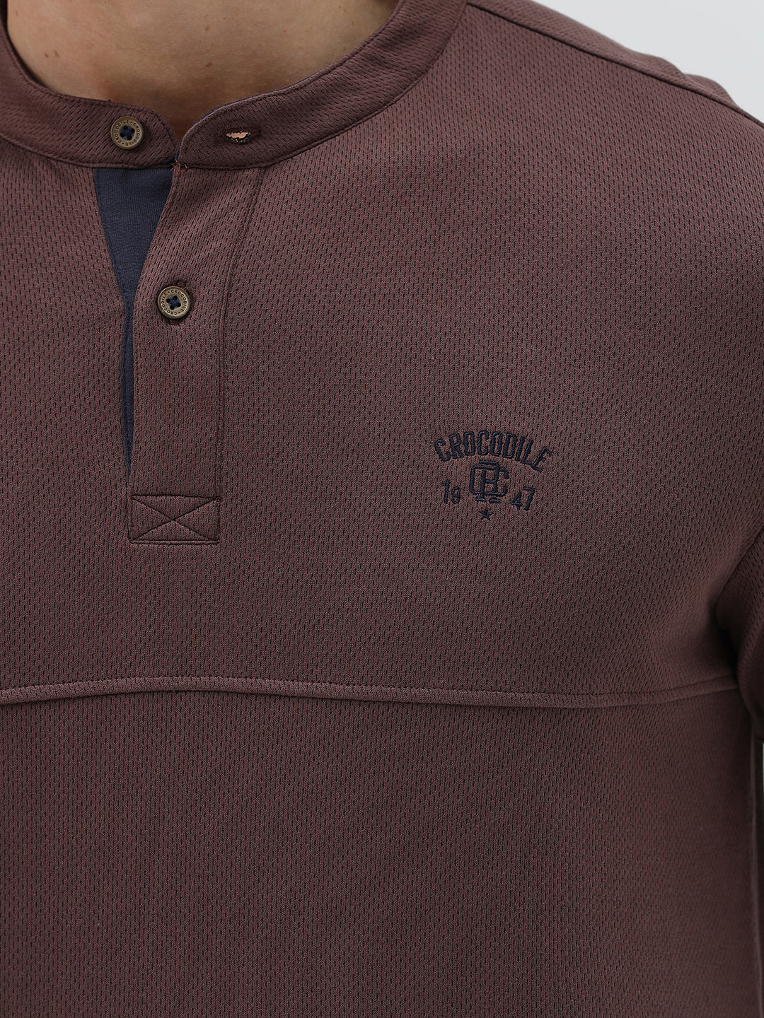 A man wearing a brown long-sleeve henley shirt with a small embroidered logo on the left chest, paired with dark blue jeans, standing against a light gray background.