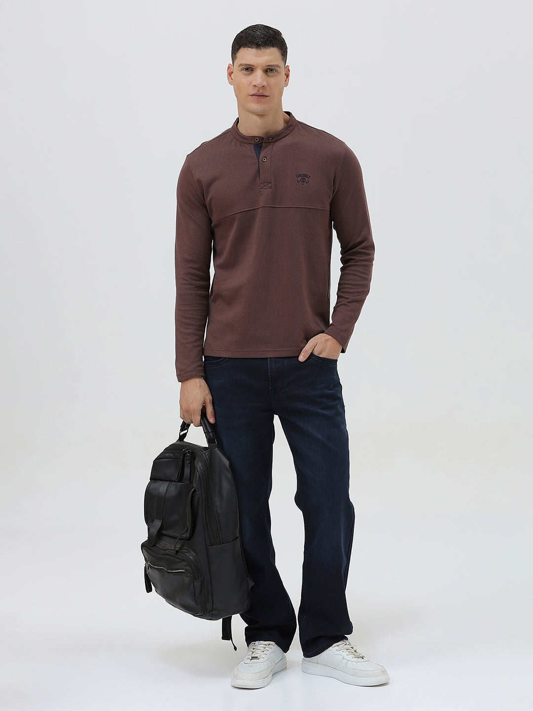 A man wearing a brown long-sleeve henley shirt with a small embroidered logo on the left chest, paired with dark blue jeans, standing against a light gray background.